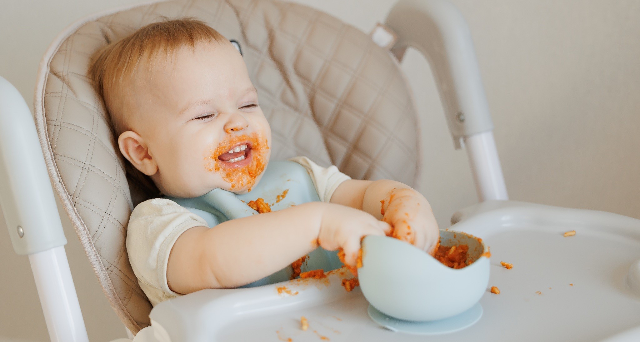 Happy baby with messy face enjoying a meal in a high chair.