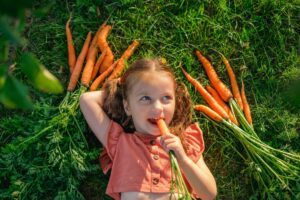 A young girl lying on grass, eating a carrot with fresh carrots beside her.