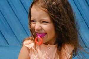 A joyful young girl smiling with a colorful pacifier in her mouth.